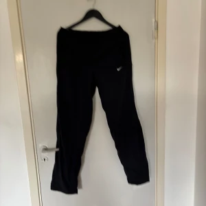 Svarta träningsbyxor från Nike - Svarta träningsbyxor från Nike med elastisk midja och meshfoder. Byxorna har raka ben, två bakfickor och en liten Nike-logga framtill. Perfekta för dig som vill ha bekväma och stilrena byxor till gymmet eller vardagsaktiviteter.