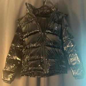 Svart pufferjacka från Trapstar - Snygg svart pufferjacka från Trapstar med glansig finish och broderad logga på ryggen. Jackan har dragkedja framtill, hög krage och rund patch med 'It's a Secret' på bröstet. Tillverkad i 100% polyester, perfekt för kalla dagar.