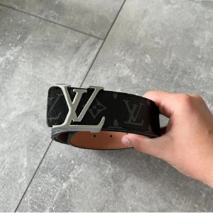Svart Louis Vuitton bälte med LV-spänne - Säljer ett svart Louis Vuitton bälte med ikoniskt LV-spänne i silver. Bältet har diskret monogrammönster och är tillverkat i exklusivt skinn. Insidan är ljusbrun och präglad med Louis Vuitton Paris made in France. Perfekt accessoar för att lyfta din outfit.
