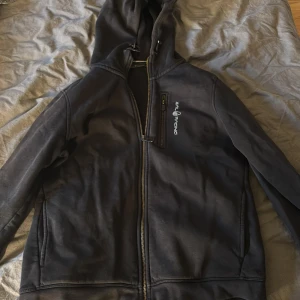 Svart fleecejacka från Sail Racing - Svart fleecejacka från Sail Racing med huva och hel dragkedja framtill. Jackan har en bröstficka med dragkedja och broderad logga på bröstet. Mjuk insida i fleece och praktiska sidfickor. Perfekt för kyliga dagar.