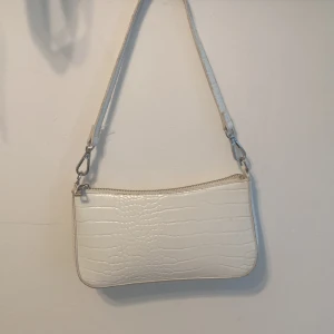 Vit axelväska i krokopräglat skinn - Snygg vit axelväska med krokodilpräglat mönster och silverfärgade metalldetaljer. Väskan har en avtagbar axelrem och stängs med dragkedja upptill. Perfekt storlek för det viktigaste och ger en clean vibe till din outfit. Axelrem:40cm väskan:22cm bred
