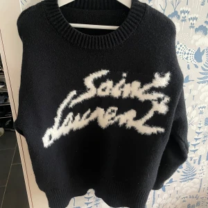 Svart stickad Saint Laurent tröja - Svart stickad tröja från Saint Laurent med vit, fluffig logga framtill i coolt skript. Tröjan har rund halsringning, ribbade muddar och en loose passform. Perfekt för dig som gillar statement-plagg och exklusiv känsla.