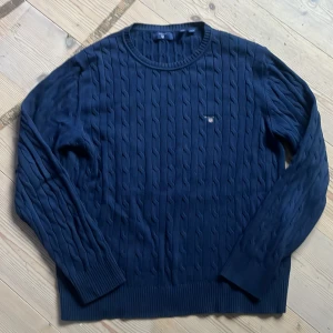 Mörkblå kabelstickad tröja från GANT - Kabelstickad mörkblå tröja från GANT med rund halsringning och broderad logga på bröstet. Tröjan har långa ärmar och ribbade muddar vid ärmslut och nederkant. Perfekt för dig som gillar klassisk och stilren look.
