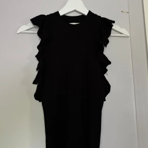 Svart ärmlös topp med volanger - Snygg svart ärmlös topp med volangdetaljer längs axlarna och sidorna. 