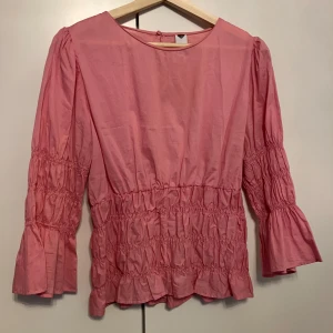 Rosa blus från Visual Clothing Project - Supersöt rosa blus med volangdetaljer och smock i midjan och på ärmarna. Blusen har rund halsringning och trekvartsärmar med lätt utsvängd avslutning. Perfekt för dig som gillar romantiska och trendiga plagg.