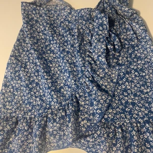 Blå blommig omlottklänning - Supersöt blå kojol med vitt blommönster och volangdetaljer. Omlottmodell som knyts i sidan, perfekt för sommarens alla tillfällen. 