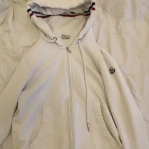 Vit zip-hoodie från Moncler - Vit hoodie från Moncler med dragkedja, huva och snörning. Klassisk logga på ärmen och snygga ränder i blått och rött på huvan. Perfekt för dig som gillar stilren look. Vid snabbköp kan priset sänkas.