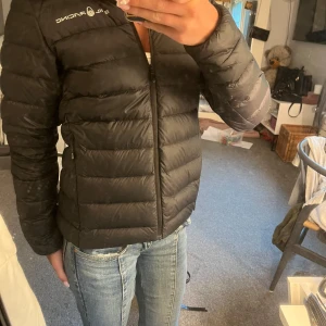 Sail Racing jacka  - Säljer en svart dunjacka från Sail Racing med stilren design och diskret logga på bröstet. Jackan är lätt, har dragkedja framtill och är fylld med 800 fill power dun för extra värme. Perfekt för kyliga dagar och enkel att matcha med allt.