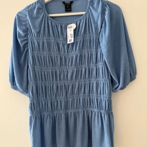 Ny Lindex blå blus, storlek S - •Ljusblå blus med smock framtill och puffärmar.  •Blusen har en rund halsringning och en lätt A-linjeformad silhuett. •Är gjord i ett mjukt, lätt material som känns skönt mot huden.  •Perfekt för dig som gillar en avslappnad men trendig stil. •Djurfritt och rökfritt hem.