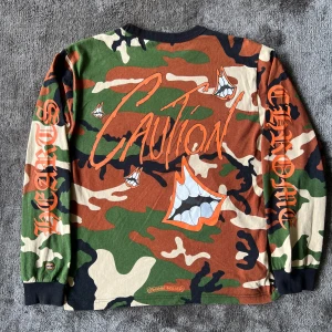 Chrome Hearts Matty boy long sleeve - Säljer en långärmad camo-tröja från Chrome Hearts med coola grafiska tryck, stora orangea CAUTION-bokstäver och tänder på både fram- och baksida. Tröjan har ficka på bröstet, svarta muddar och tryck på ärmarna. 