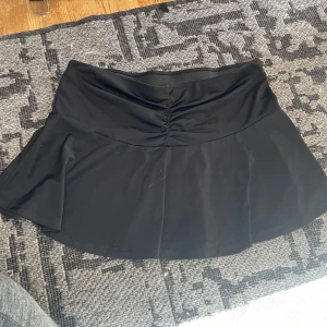 Svart kortkjol från H&M Divided - Svart kortkjol från H&M Divided med snygg rynkdetalj framtill i midjan. Kjolen är lätt utsvängd och har en bekväm resår i midjan. Tillverkad i stretchig polyesterblandning som sitter skönt och följer kroppen.