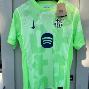 Barça fotbollströja - Snygg FC Barcelona fotbollströja från Nike i neonljusgrön färg med mörkblå detaljer och klubbmärke på bröstet. Tröjan har korta ärmar, modern passform och är tillverkad i ett lätt, ventilerande funktionsmaterial. Perfekt för dig som vill sticka ut på planen.