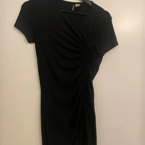 Svart kortklänning från H&M - Svart kortklänning från H&M med rynkdetalj längs ena sidan. Klänningen har rund halsringning och korta ärmar. Tight passform men stretchig Knappt använd! 