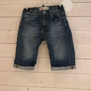 Levi's 511 jeansshorts slim fit - Snygga jeansshorts från Levi's modell 511 i slim fit. Klassisk blå tvätt med slitningar och uppvikta benslut. Femficksmodell med Levi's ikoniska läderpatch bak och röda logotag. Perfekta för en avslappnad stil under varma dagar.  Size 14 år 