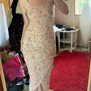 Ljusblommig långklänning med drapering - Supersnygg långklänning i ljusa toner med blommigt mönster och draperad effekt längs hela kroppen. Klänningen har breda axelband, fyrkantig ringning och omlott-detalj nertill. Perfekt för sommarens alla tillfällen!