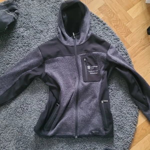 Svart och grå hooded jacka Grounded - Mörkgrå och svart jacka med huva från Grounded. Jackan har hel dragkedja, tre fickor med dragkedja och tryckt logga på bröstet. Materialet är mjukt och stretchigt, perfekt för chill eller utomhus. Snygg passform och sportig look.