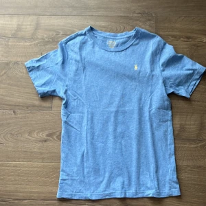 Blå t-shirt från Polo Ralph Lauren - Snygg blå t-shirt från Polo Ralph Lauren med klassisk gul broderad logga på bröstet. T-shirten har rund halsringning och korta ärmar. Tillverkad i mjuk bomull som är skön mot huden. Perfekt för en chill och stilren look.