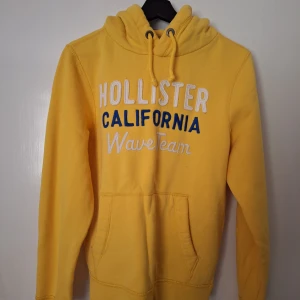 Gul Hollister hoodie - Gul hoodie från Hollister med broderad text i vitt och blått framtill. Klassisk känguruficka, snörning i huvan och mjukt material. Perfekt för dig som vill sticka ut med färg och hålla dig bekväm. Snygg till jeans eller joggers.