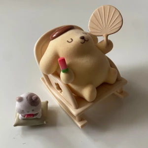 Sanrio pompompurin figur - Söt pompompurin figur, en större hundliknande karaktär som sitter i en gungstol och håller en solfjäder och en glass eller vattenmelon, samt en mindre kattliknande figur på en kudde med en bit vattenmelon. Figurerna är ca 7 cm höga och har en mjuk, pastellfärgad design. 