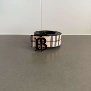 Rutig Burberry bälte med B-spänne - Snyggt bälte från Burberry med klassiskt rutigt mönster i beige, svart och rött. Bältet har ett stort svart B-spänne framtill och är gjort i skinn med tygdetaljer. Perfekt accessoar för att lyfta din outfit med en lyxig touch.