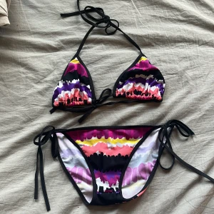 Färgglad bikini med knyt i sidan - Snygg bikini med färgstarkt mönster i svart, lila, rosa, vit, gul och röd. Triangeltopp med knyt i nacken och ryggen samt matchande trosa med knyt i sidorna. Perfekt för stranden och sommarens alla bad. Materialet är mjukt och stretchigt syntet. ALDRIG ANVÄND
