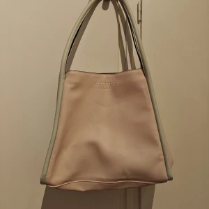 Ljusrosa axelväska från Victoria's Secret - Stilren och rymlig axelväska från Victoria's Secret i ljusrosa med vitgrå detaljer och handtag. Väskan är tillverkad i fakeläder med slät yta och har ett stort huvudfack med tryckknappsstängning. Perfekt för dig som vill ha en enkel men snygg väska till vardagen.