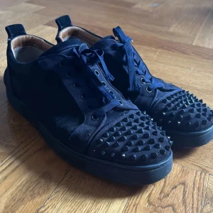 Svarta Christian Louboutin söker byte  - Snygga svarta sneakers med coola svarta nitar över tån och mockadetaljer. Skorna har snörning och en platt sula, perfekt för dig som vill sticka ut med en edgy stil. Bekväma och trendiga med en unik look.