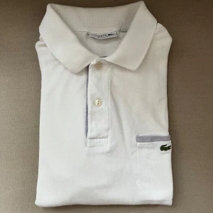 Vit pikétröja från Lacoste - Klassisk vit pikétröja från Lacoste i bomull med broderad krokodil på bröstfickan. Tröjan har krage, knäppning med två knappar och diskreta grå detaljer vid fickan. Perfekt för en clean och sportig stil. 