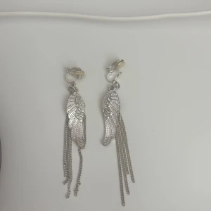 Silvriga clips örhängen med vingar och kedjor - Snygga silvriga clips örhängen med form av vingar och glittrande stenar. Hängande kedjor ger extra edge och cool känsla. Perfekta för dig som vill sticka ut med ett unikt smycke.