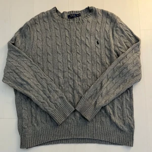 Grå kabelstickad tröja Polo Ralph Lauren - Kabelstickad grå tröja från Polo Ralph Lauren i klassisk modell med rund hals och broderad logga på bröstet.