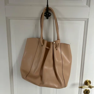 Beige shoppingväska i skinn - Stilren beige shoppingväska i mjukt skinn med långa handtag och enkel knappstängning. Väskan har ett rymligt huvudfack och en innerficka med dragkedja för småsaker. Perfekt för dig som vill ha plats för allt du behöver under dagen.
