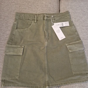 Grön cargokjol i denim, stl 38 - Snygg cargokjol i denim med coola fickor på sidorna och bälteshällor. Kjolen är i en dämpad grön färg och har en rak, kort passform