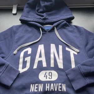Marinblå hoodie från GANT XS - Snygg marinblå hoodie från GANT i storlek XS med stora vita tryck på bröstet där det står 'GANT 49 NEW HAVEN'. Tröjan har huva med grå snören och långa ärmar. Passar även mindre storlekar än xs, dvs liten i storleken. Perfekt för att bygga upp en snygg stil!