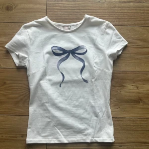 Vit t-shirt med rosett från Nelly - Vit t-shirt från Nelly. Aldrig använd och är i storlek Xs. Den är väldigt fin men lite kort i ärmen.