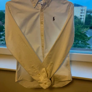 Vit skjorta från Ralph Lauren slim fit - Klassisk vit skjorta från Ralph Lauren i slim fit-modell. Skjortan är tillverkad i 100% bomull och har knappar framtill samt det ikoniska broderade polospelar-loggan på bröstet. Perfekt för en clean och stilren look.