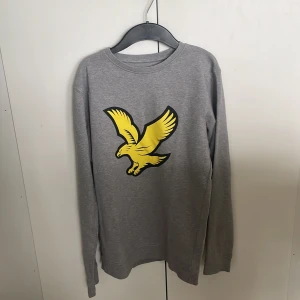 Grå Lyle & Scott tröja med gul örn - Snygg grå långärmad tröja från Lyle & Scott med stort gult örntryck på bröstet. Klassisk rund halsringning och mjukt material som känns skönt mot huden. Perfekt för dig som gillar stilrena plagg med en cool detalj framtill. Passar som XS