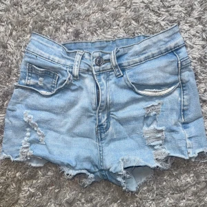 Ljusblå slitna jeansshorts - Snygga ljusblå jeansshorts med slitna detaljer och fransig kant. Klassisk femficksmodell med dragkedja och knapp framtill. Perfekta för varma dagar när du vill ha en avslappnad och trendig look.