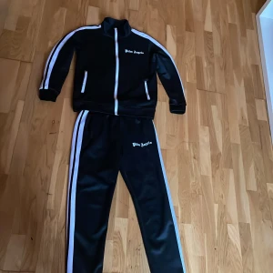 Palm Angels svart track jacket - Svart track jacket från Palm Angels med vita ränder längs ärmarna och dragkedja framtill. Jackan har hög krage, två fickor med dragkedja och logga på bröstet. Perfekt för en sportig och trendig look.