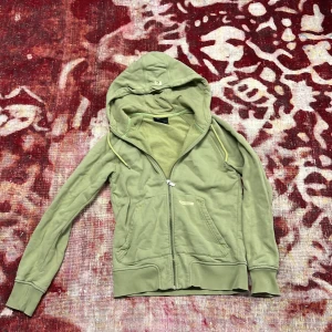 Grön hoodie från Peak Performance M - Snygg ljusgrön hoodie från Peak Performance med dragkedja och stor huva. Tröjan har ett unikt tryck och broderi på ryggen, samt praktiska fickor framtill. Mjuk insida i fleece och coola detaljer som snörning i huvan. Perfekt för chill dagar eller till träning. Ett litet hål på ryggen