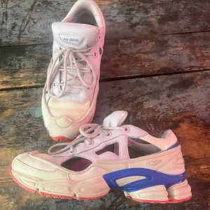 Raf Simons x Adidas Ozweego sneakers - Unika Raf Simons x Adidas Ozweego sneakers i ljusbeige och vitt med detaljer i blått och rött. Chunky sula, mesh och skinnpaneler, coola cut-outs på sidorna och snörning framtill. Ett par tillhörande strumpor medföljer, oanvända