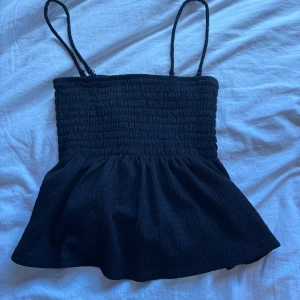 Svart smockad linnetopp med volang - Supersöt svart linnetopp med smockad överdel och volang nertill. Toppen har avtagbara tunna axelband så du kan bära den både som axelbandslös och med band. Perfekt till jeans eller kjol för en chill look.