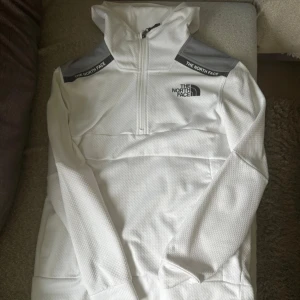 Vit hoodie från The North Face M - Snygg vit hoodie från The North Face med grå axelpartier och logga på bröstet. Tröjan har huva, lång ärm och half zip-dragkedja framtill. Perfekt för dig som gillar sportig och clean stil. Storlek M