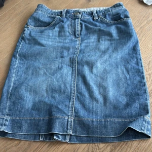 Jeanskjol från GANT med slits bak - Klassisk blå jeanskjol från GANT med femficksmodell, bälteshällor och snygg slits bak. Kjolen har dragkedja och knapp framtill samt tydliga sömmar. Perfekt för dig som gillar en tidlös och avslappnad stil.