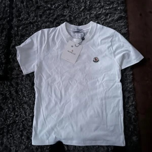 Vit Moncler t-shirt i bomull - Snygg vit t-shirt från Moncler med klassisk rund hals och liten broderad logga på bröstet. Tillverkad i mjuk bomull och har en enkel, clean look som passar perfekt till jeans eller shorts.
