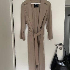 Beige lång trenchcoat från Zara  - Helt ny beige lång trenchcoat med knytning från Zara. Tunt tyg. Aldrig använd. Oversize modell så passar även S,M beroende på hur man vill att den sitter. Ca 122 cm lång från krage och neråt.
