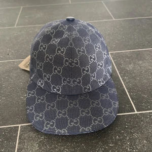 Blå keps från Gucci med GG-mönster - Snygg blå keps från Gucci med klassiskt GG-mönster över hela kepsen. Justerbar rem baktill i blått skinn och böjd skärm. Tillverkad i bomullsmix med exklusiv känsla. Perfekt för dig som vill sticka ut med en lyxig accessoar.