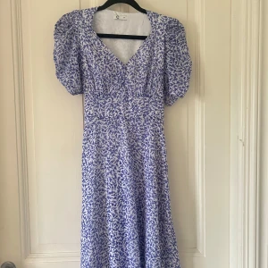 Blåvit blommig klänning med puffärm - Supersöt blåvit klänning med blommigt mönster, puffiga korta ärmar och v-ringning. Klänningen har knytband i midjan och en lätt utställd kjol som ger en romantisk vibe. Perfekt för varma dagar när du vill känna dig extra fin.