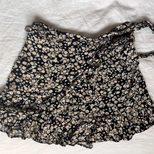 Svart blommig omlottkjol med volang - Supersöt svart kort omlottkjol med vita och gula blommor. Kjolen har volangkant nertill och knyts i sidan för en snygg passform. Perfekt för dig som gillar en feminin och lekfull stil.💞 Storlek är barn L men tjolen är justerbar alltså passar XS, S och M i vuxenstorleckar😊