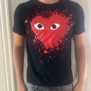 Svart Comme des Garçons Play t-shirt - Svart t-shirt från Comme des Garçons Play med ikoniskt rött hjärta med ögon och färgstänk på bröstet.