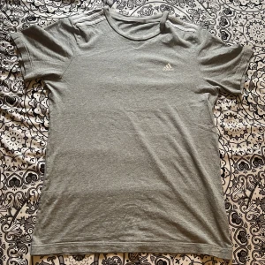 Grå t-shirt från Adidas - Snygg grå t-shirt från Adidas med klassiska tre ränder på axlarna och liten logga på bröstet. T-shirten är i mjuk bomull och har en enkel, clean look med rund hals och korta ärmar. Perfekt för en sportig vibe eller chill vardagsstil.